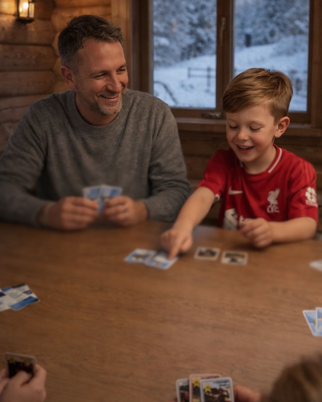 Pappa och son spelar kort vid ett träbord i en timmerstuga med snö utanför fönstret, ytterligare två spelare syns i bildens nederkant
